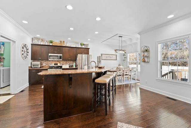 kitchen featuring stainless steel appliances, a center island with sink, dark brown cabinetry, decorative light fixtures, and dark hardwood / wood-style flooring