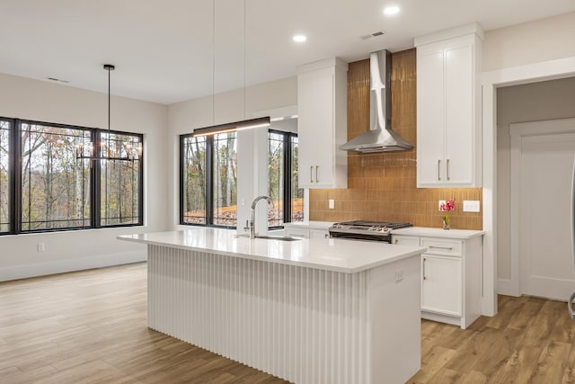 kitchen featuring stove, an island with sink, decorative light fixtures, and wall chimney range hood