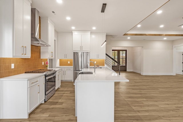 kitchen featuring a kitchen island with sink, white cabinets, wall chimney range hood, decorative light fixtures, and stainless steel appliances