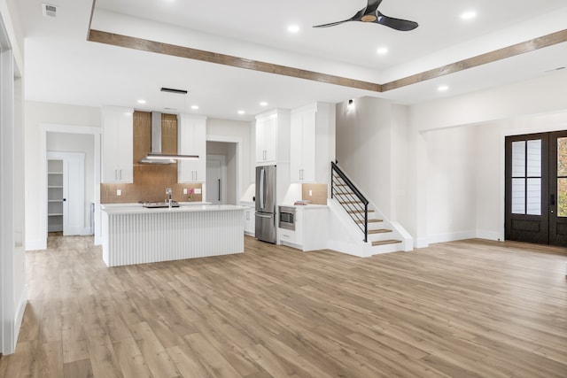 kitchen featuring wall chimney exhaust hood, tasteful backsplash, a kitchen island with sink, white cabinets, and appliances with stainless steel finishes