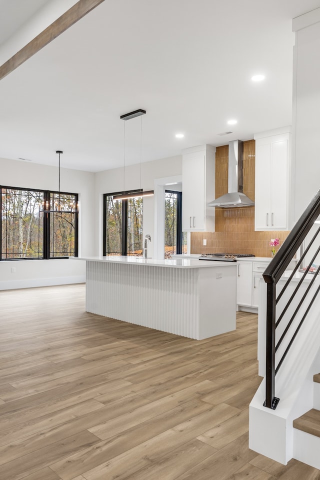 kitchen with wall chimney range hood, a notable chandelier, decorative light fixtures, a center island with sink, and white cabinets