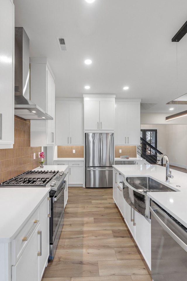 kitchen with sink, hanging light fixtures, wall chimney exhaust hood, appliances with stainless steel finishes, and white cabinetry