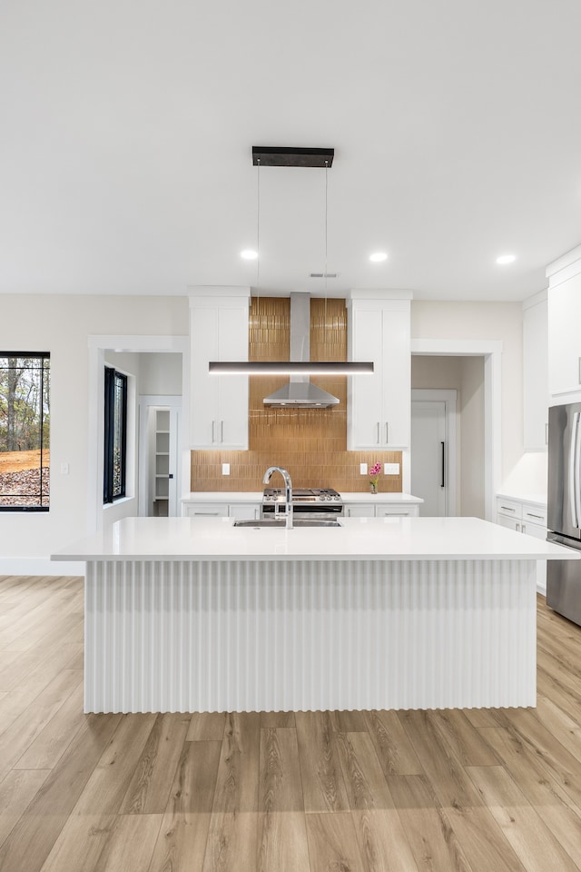 kitchen with stainless steel fridge, wall chimney exhaust hood, pendant lighting, a center island with sink, and white cabinetry