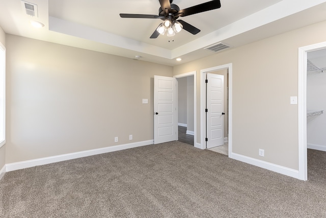 unfurnished bedroom with a walk in closet, a tray ceiling, a closet, ceiling fan, and carpet