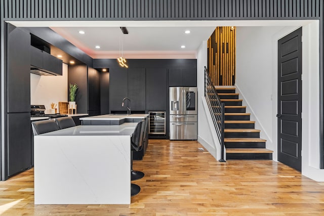 kitchen with an island with sink, hanging light fixtures, wine cooler, light wood-type flooring, and appliances with stainless steel finishes