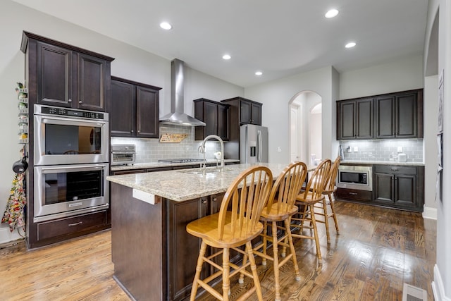 kitchen with wall chimney exhaust hood, an island with sink, wood-type flooring, a kitchen bar, and appliances with stainless steel finishes