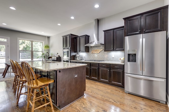 kitchen featuring an island with sink, appliances with stainless steel finishes, light wood-type flooring, wall chimney exhaust hood, and sink