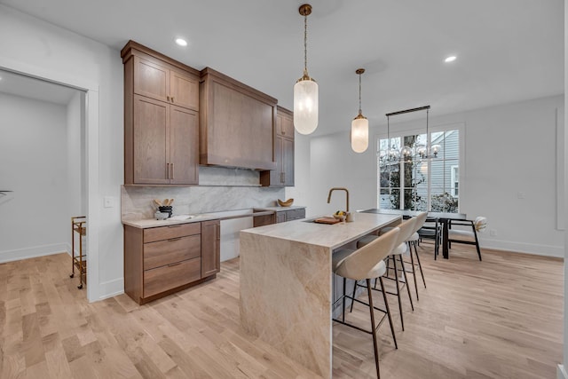 kitchen with sink, hanging light fixtures, backsplash, an island with sink, and light wood-type flooring