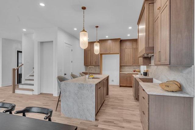 kitchen featuring sink, backsplash, hanging light fixtures, a kitchen island with sink, and light wood-type flooring