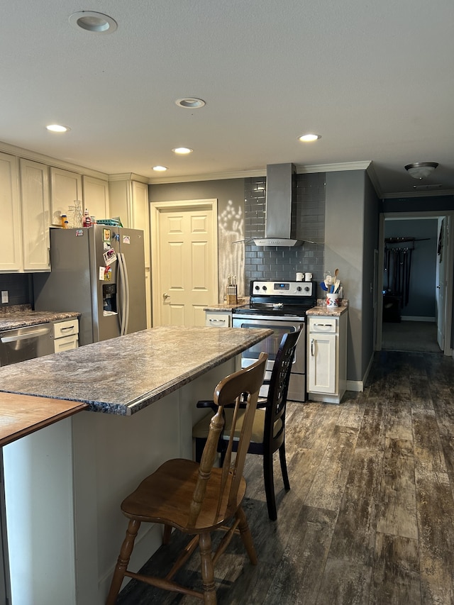 kitchen featuring wall chimney range hood, appliances with stainless steel finishes, dark wood-type flooring, ornamental molding, and a breakfast bar area