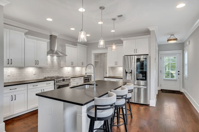 kitchen featuring a kitchen island with sink, wall chimney range hood, stainless steel appliances, pendant lighting, and white cabinets