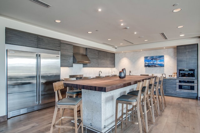 kitchen with light hardwood / wood-style flooring, wall chimney exhaust hood, built in fridge, and butcher block countertops