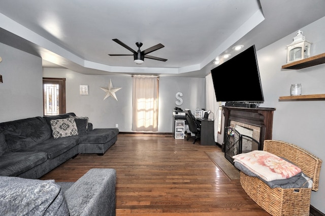 living room featuring ceiling fan, a stone fireplace, a tray ceiling, and dark hardwood / wood-style floors