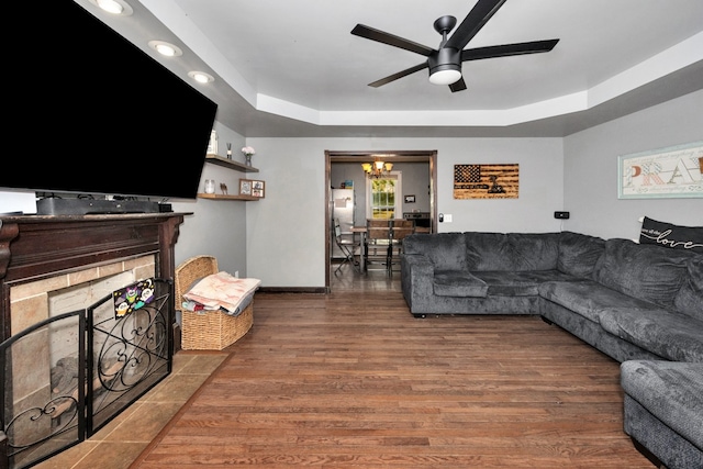 living room with a stone fireplace, ceiling fan with notable chandelier, a raised ceiling, and dark hardwood / wood-style flooring