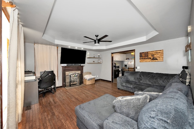 living room with ceiling fan, a tray ceiling, a fireplace, and dark hardwood / wood-style flooring