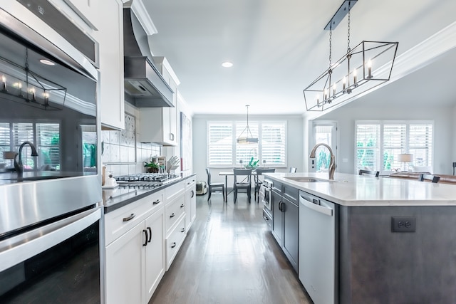 kitchen featuring wall chimney exhaust hood, stainless steel appliances, white cabinets, hanging light fixtures, and an island with sink