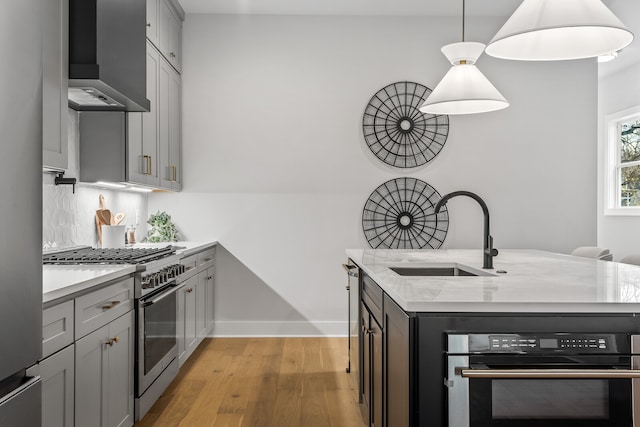 kitchen featuring wall chimney range hood, light stone counters, stainless steel appliances, light hardwood / wood-style floors, and sink