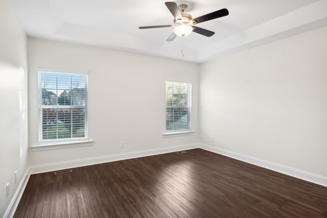 empty room with a raised ceiling, plenty of natural light, dark wood-type flooring, and ceiling fan