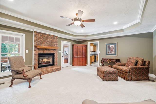 carpeted living room featuring ornamental molding, a textured ceiling, a brick fireplace, a tray ceiling, and ceiling fan