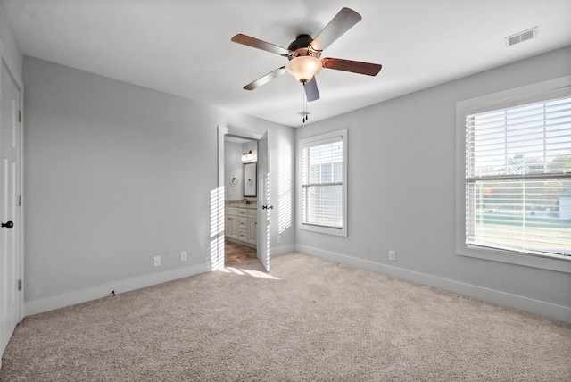 unfurnished bedroom with ensuite bath, multiple windows, light colored carpet, and ceiling fan