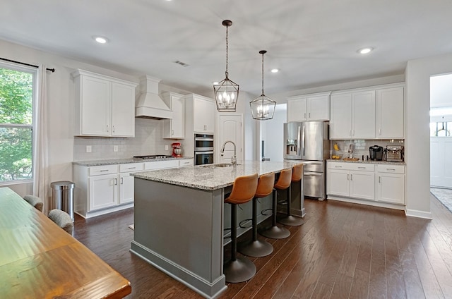 kitchen featuring appliances with stainless steel finishes, custom exhaust hood, white cabinetry, and a center island with sink