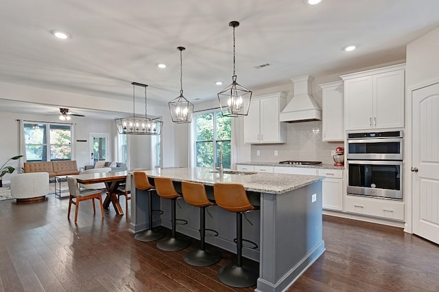 kitchen featuring stainless steel double oven, white cabinetry, pendant lighting, custom range hood, and a kitchen island with sink