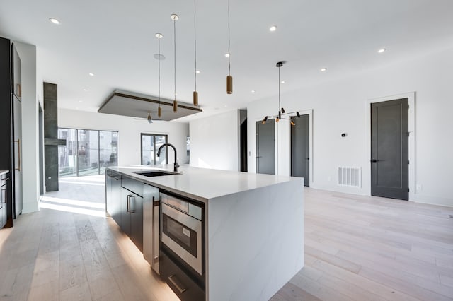 kitchen with a large island with sink, stainless steel oven, sink, decorative light fixtures, and light hardwood / wood-style floors