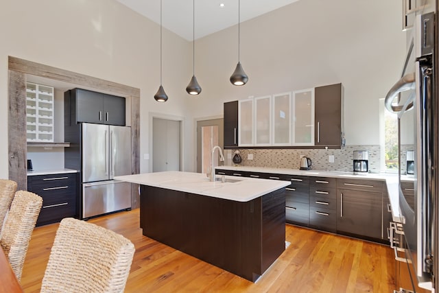 kitchen featuring light wood-type flooring, a towering ceiling, sink, a center island with sink, and stainless steel refrigerator