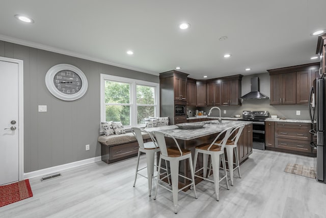 kitchen with light stone countertops, appliances with stainless steel finishes, light hardwood / wood-style floors, wall chimney exhaust hood, and a center island with sink