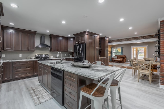 kitchen featuring black appliances, sink, light hardwood / wood-style floors, wall chimney exhaust hood, and a center island with sink