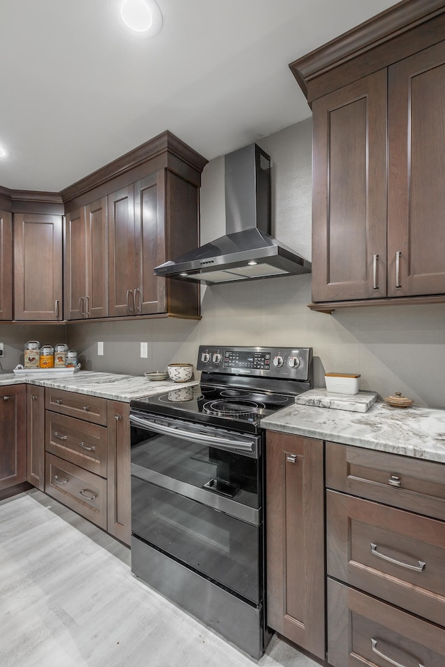 kitchen featuring black range with electric cooktop, light wood-type flooring, wall chimney exhaust hood, dark brown cabinetry, and light stone counters