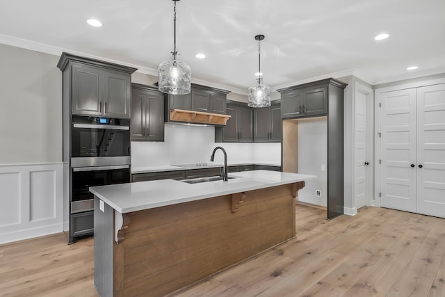 kitchen featuring pendant lighting, crown molding, an island with sink, double oven, and light hardwood / wood-style floors