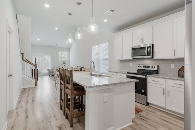 kitchen with white cabinets, an island with sink, light wood-type flooring, decorative light fixtures, and stainless steel appliances