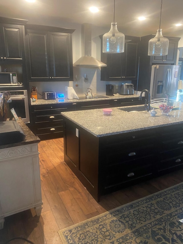 kitchen with a center island with sink, appliances with stainless steel finishes, light wood-type flooring, wall chimney exhaust hood, and sink