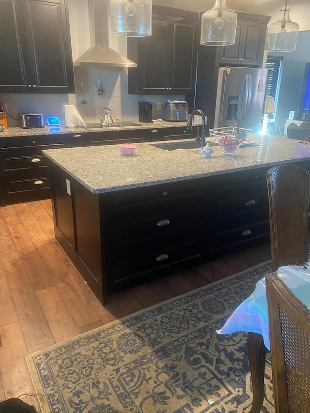 kitchen with wall chimney range hood, a center island with sink, stainless steel fridge with ice dispenser, light hardwood / wood-style flooring, and black electric cooktop