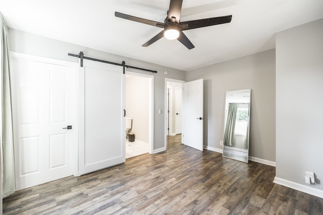 unfurnished bedroom featuring ensuite bathroom, dark hardwood / wood-style floors, a barn door, and ceiling fan