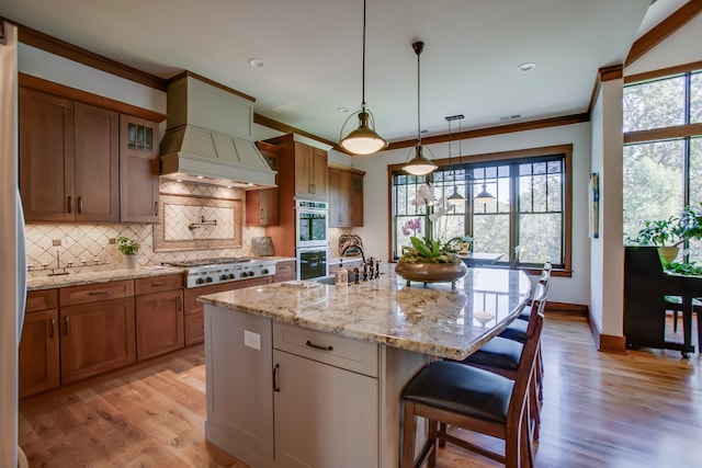 kitchen featuring light stone counters, an island with sink, decorative light fixtures, appliances with stainless steel finishes, and custom exhaust hood