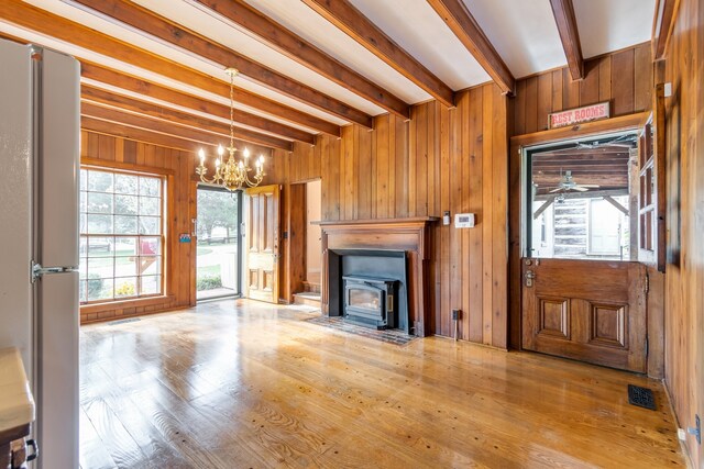 unfurnished living room with beam ceiling, wooden walls, ceiling fan with notable chandelier, and light wood-type flooring