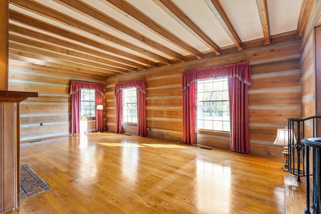 unfurnished living room featuring wooden walls, plenty of natural light, beamed ceiling, and light hardwood / wood-style floors