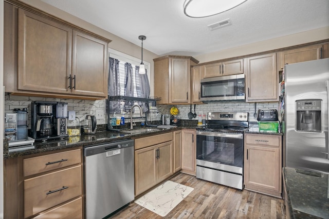 kitchen featuring appliances with stainless steel finishes, dark stone counters, sink, light hardwood / wood-style flooring, and hanging light fixtures