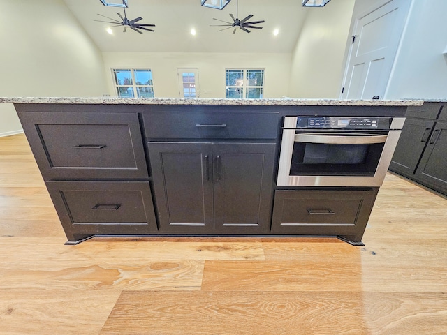 kitchen with stainless steel oven, vaulted ceiling, light stone counters, light hardwood / wood-style floors, and ceiling fan