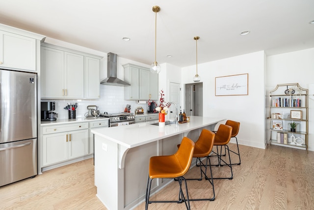 kitchen with wall chimney range hood, backsplash, light hardwood / wood-style floors, a center island with sink, and appliances with stainless steel finishes