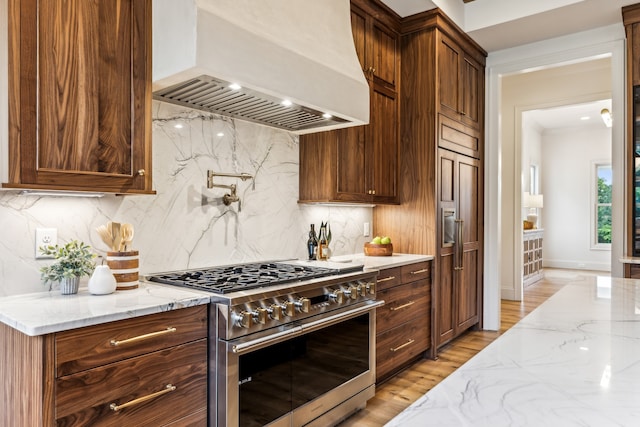 kitchen featuring light stone countertops, backsplash, light hardwood / wood-style floors, stainless steel range, and premium range hood