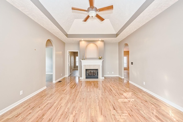 unfurnished living room with a tile fireplace, light wood-type flooring, ceiling fan, a raised ceiling, and a textured ceiling