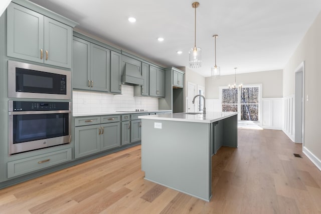 kitchen with built in microwave, stainless steel oven, custom range hood, pendant lighting, and a kitchen island with sink