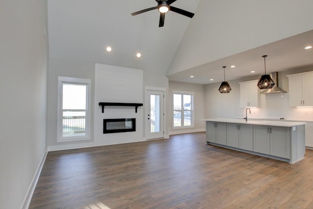 kitchen with pendant lighting, wall chimney range hood, a kitchen island with sink, white cabinetry, and high vaulted ceiling