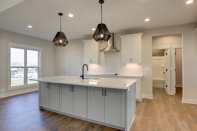 kitchen featuring sink, wall chimney range hood, white cabinetry, light hardwood / wood-style floors, and a center island with sink