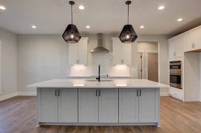 kitchen with white cabinets, hardwood / wood-style flooring, a kitchen island with sink, stainless steel double oven, and wall chimney exhaust hood