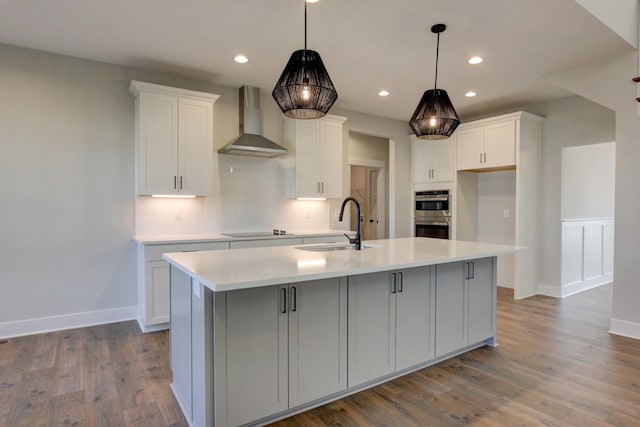 kitchen featuring white cabinetry, black electric stovetop, a center island with sink, decorative light fixtures, and wall chimney exhaust hood