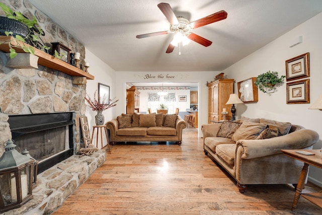 living room with a stone fireplace, light wood-type flooring, a textured ceiling, and ceiling fan